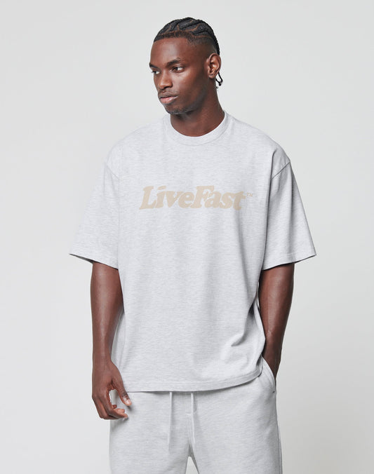A man wears the LIVE FAST DIE YOUNG Basic Tee in oversize fit, light gray with a beige front print, paired with matching gray shorts. He stands against a plain light background, looking left with one hand in his pocket.
