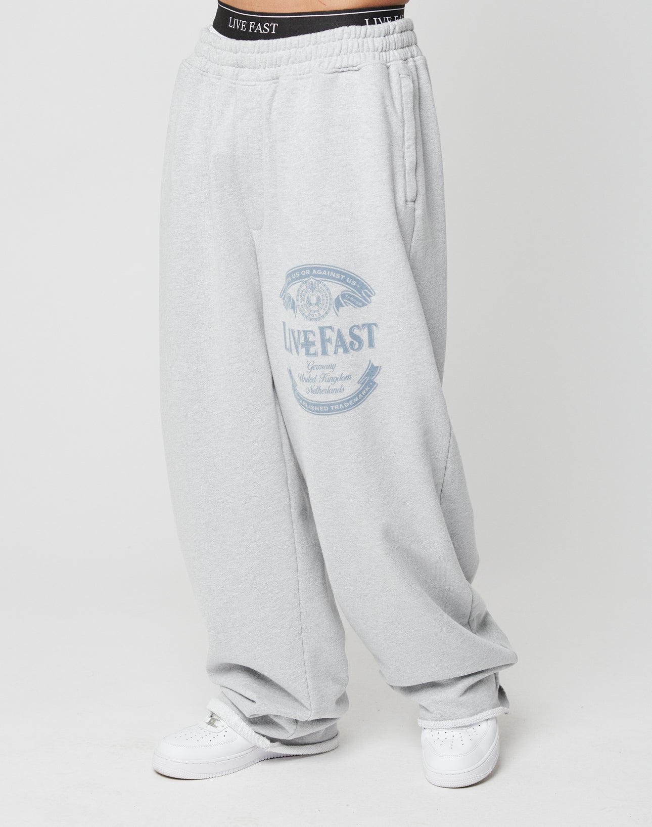Person wearing Loyal Wide Jogger by LIVE FAST DIE YOUNG—loose-fitting light gray sweatpants with oversize fit, wide leg, elastic waistband, side pockets, and LIVE FAST graphic on the upper thigh. Paired with white sneakers.