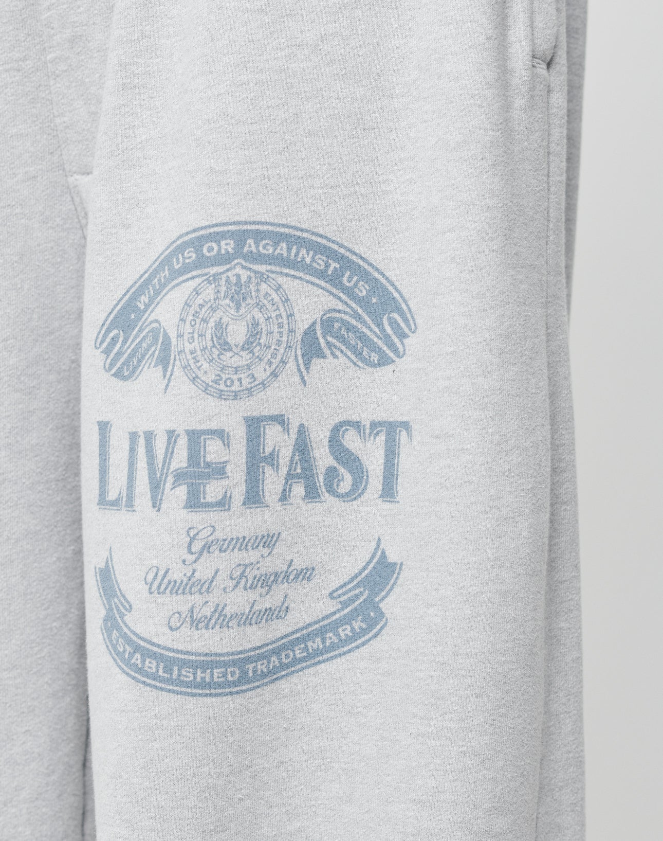The Loyal Wide Jogger by LIVE FAST DIE YOUNG features light gray fabric with a blue LIVE FAST print, banners for Germany, UK, Netherlands, and Established Trademark. It offers an oversize fit and elastic waistband for extra comfort.
