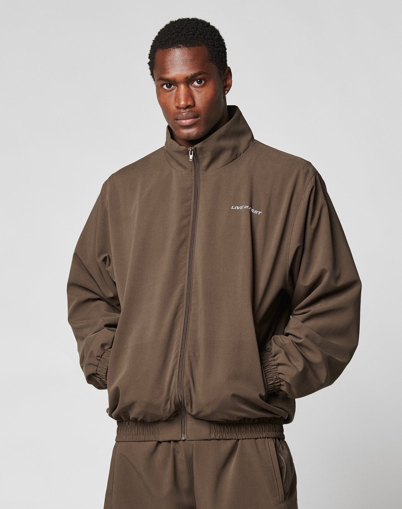 A man faces the camera with hands in pockets, wearing the LFDY Trackjacket by LIVE FAST DIE YOUNG—an oversize fit brown zip-up polyester jacket with a small white logo and reflective print on the chest. The background is plain and light-colored.