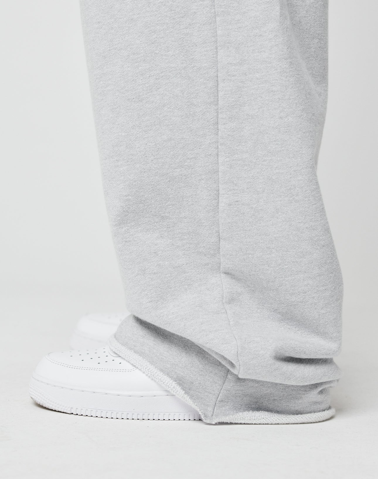 A close-up side view shows a person wearing LIVE FAST DIE YOUNG Loyal Wide Jogger in light gray with an elastic waistband and wide leg, paired with white sneakers. The joggers slightly cover the shoe tops against a plain light background.