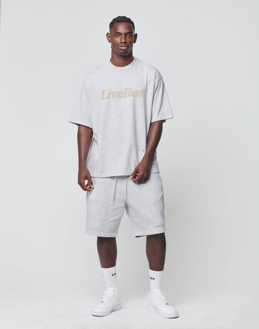 A man faces forward against a plain backdrop, wearing a gray oversized LIVE FAST DIE YOUNG Logo-Print t-shirt, matching Basic Heavy Shorts with an elastic waistband, white crew socks, and white sneakers.