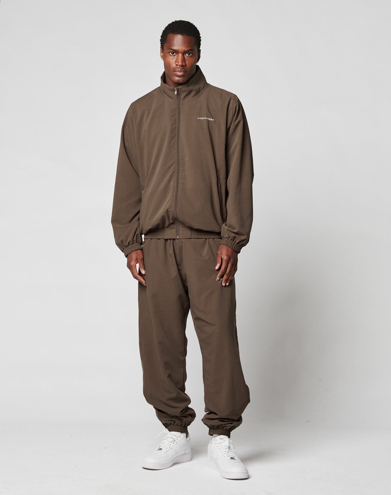 A man stands against a plain background wearing the LIVE FAST DIE YOUNG LFDY Trackjacket and matching pants, styled in an oversize fit with white sneakers. He faces forward neutrally, arms relaxed at his sides.