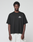 A young man with short curly hair wears the LIVE FAST DIE YOUNG LFDY Real Ones Tee in Oversize Fit with white chest text and white shorts, standing against a plain light gray background.