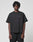 A person with short dreadlocks wears the LIVE FAST DIE YOUNG Basic Trademark Tee in an oversize fit with black pants, both made from 100% Baumwolle, standing against a plain light gray background facing the camera neutrally.