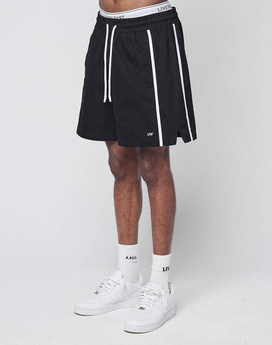 A person wearing LIVE FAST DIE YOUNG Basic Cotton Shorts—black with white vertical stripes, elastic waistband, regular fit—paired with white socks and sneakers, stands against a plain white background.