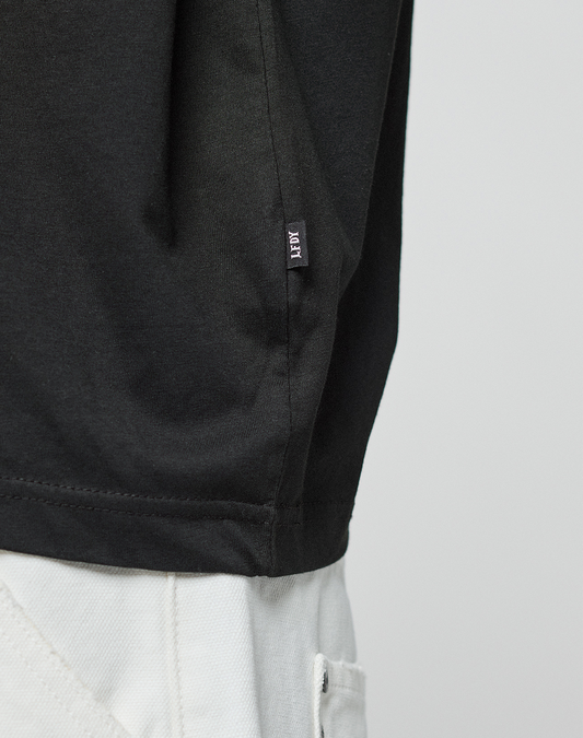 Close-up of a person wearing the LFDY Real Ones Tee by LIVE FAST DIE YOUNG in black, made from 100% Baumwolle, paired with light pants. The lower shirt features a small black tag with white text against a plain background.