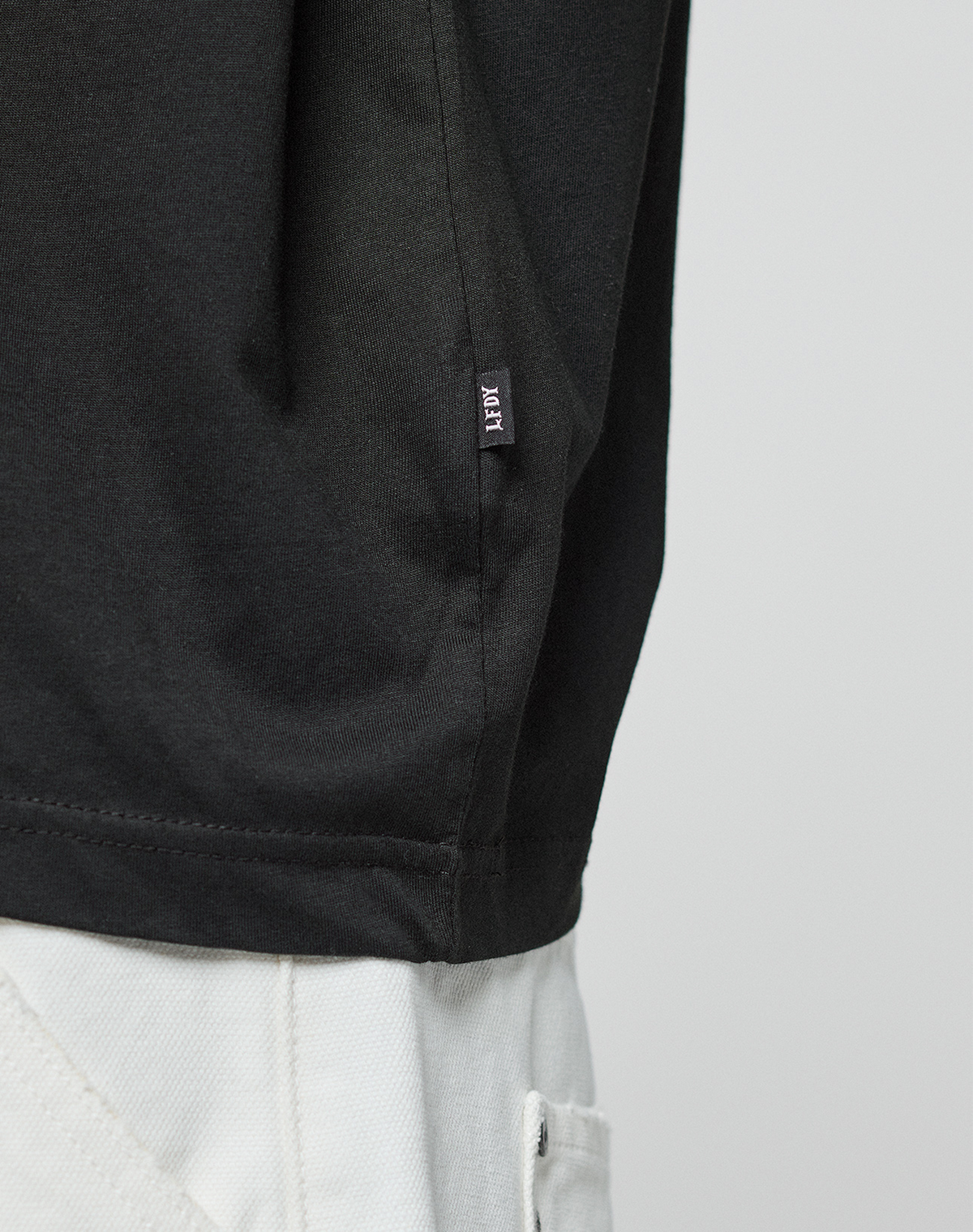 Close-up of a person wearing the LFDY Real Ones Tee by LIVE FAST DIE YOUNG in black, made from 100% Baumwolle, paired with light pants. The lower shirt features a small black tag with white text against a plain background.