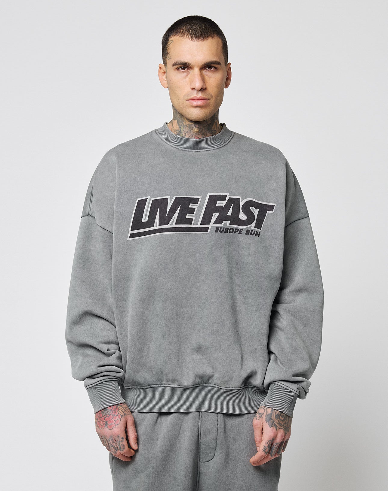 A man with short dark hair and hand tattoos faces forward, wearing the LIVE FAST DIE YOUNG Live Fast Europe Run Sweater in oversize fit with a bold frontprint. The background is plain white.
