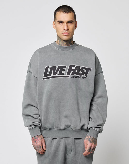 A man with short dark hair and hand tattoos faces forward, wearing the LIVE FAST DIE YOUNG Live Fast Europe Run Sweater in oversize fit with a bold frontprint. The background is plain white.