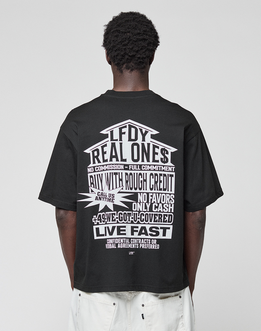 A person with short curly hair, seen from behind, wears the LFDY Real Ones Tee by LIVE FAST DIE YOUNG—a loose black 100% Baumwolle T-shirt with bold white "Real Ones" graphic—paired with loose white shorts.