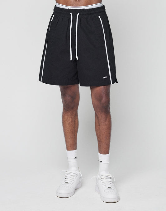 A person wears LIVE FAST DIE YOUNG Basic Cotton Shorts—black with white vertical stripes, logo print, elastic waistband, drawstring, and side pockets—plus white crew socks and sneakers. Their upper body isn’t visible against a light background.