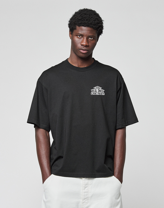 A person wears the LIVE FAST DIE YOUNG LFDY Real Ones Tee, a loose black T-shirt with front and back print, and white pants, standing against a plain light gray background with hands in pockets, facing the camera.