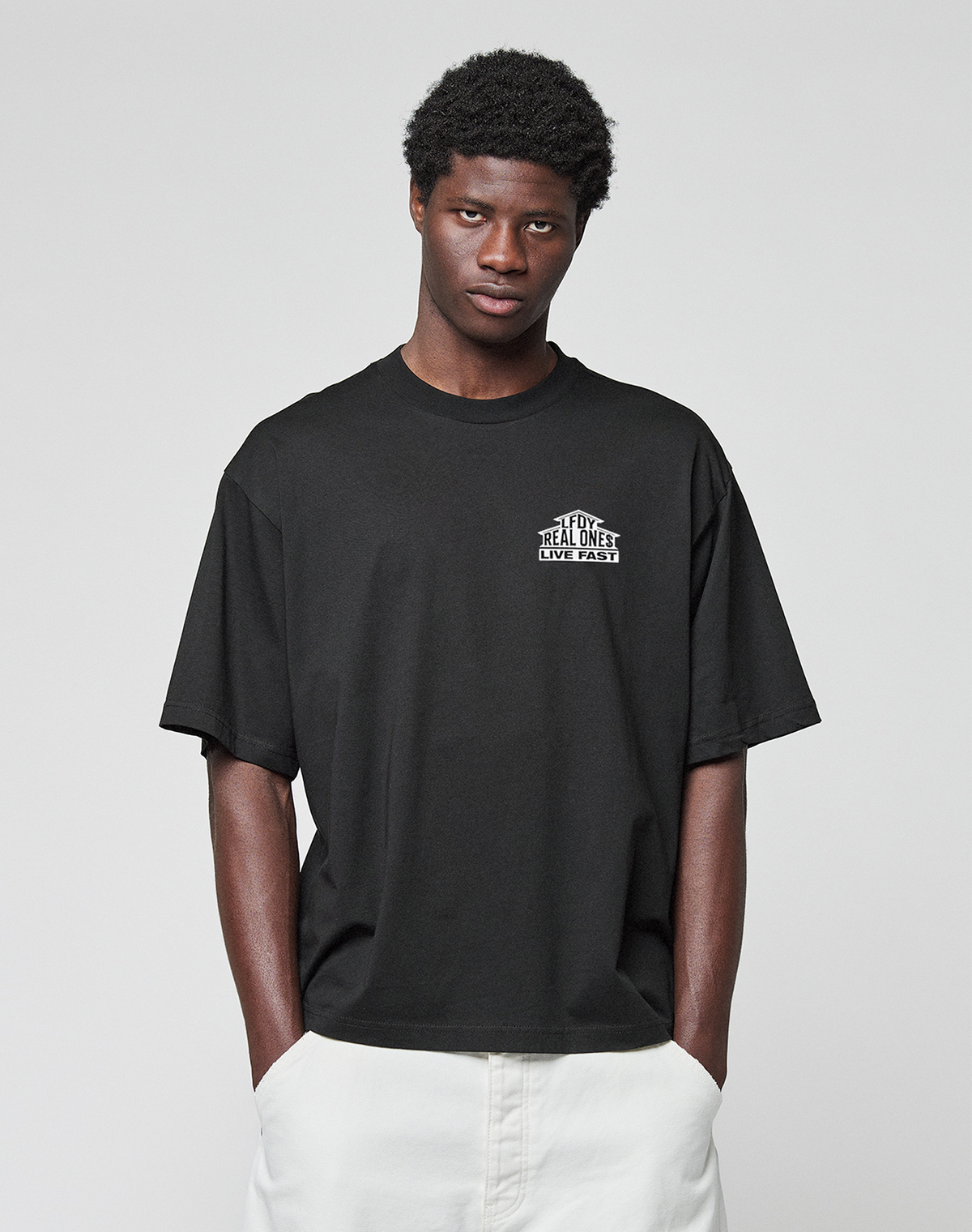 A person wears the LIVE FAST DIE YOUNG LFDY Real Ones Tee, a loose black T-shirt with front and back print, and white pants, standing against a plain light gray background with hands in pockets, facing the camera.