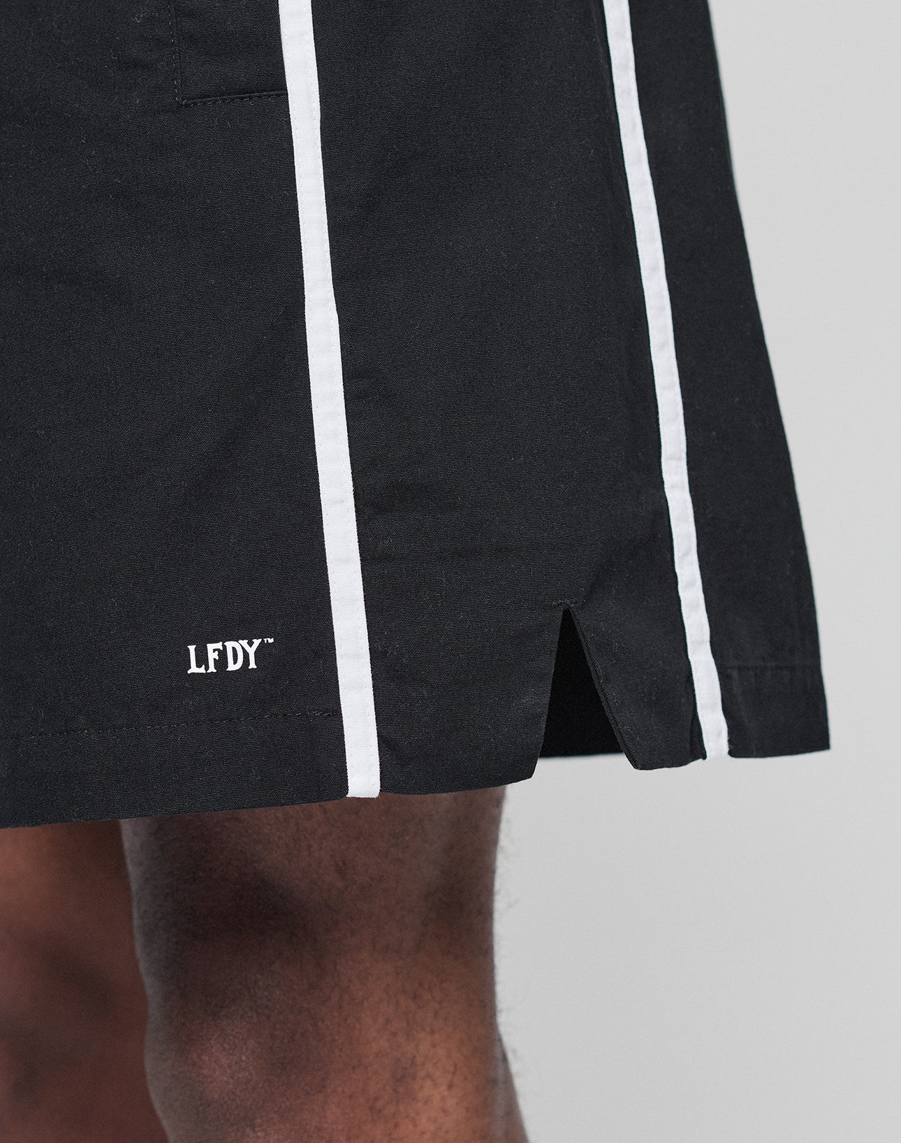 Close-up of a person wearing Basic Cotton Shorts by LIVE FAST DIE YOUNG, featuring a regular fit, black color with white vertical stripes, elastic waistband, front slit, and white LFDY logo print near the above-the-knee hem.