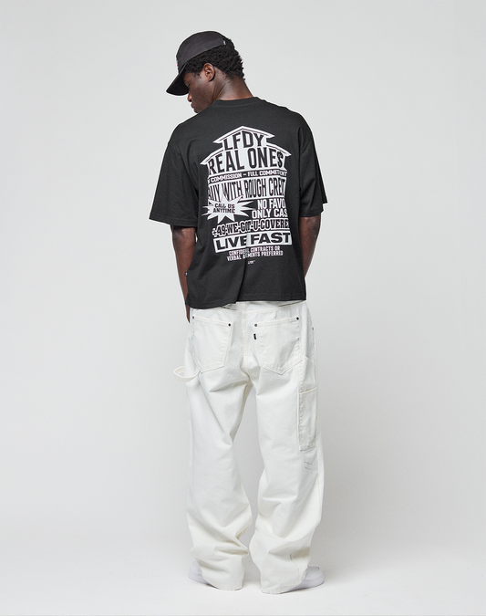 A person faces away, wearing a black LIVE FAST DIE YOUNG LFDY Real Ones Tee made of 100% Baumwolle with bold white front and back prints, a black cap, and loose-fitting white pants, set against a plain light grey background.