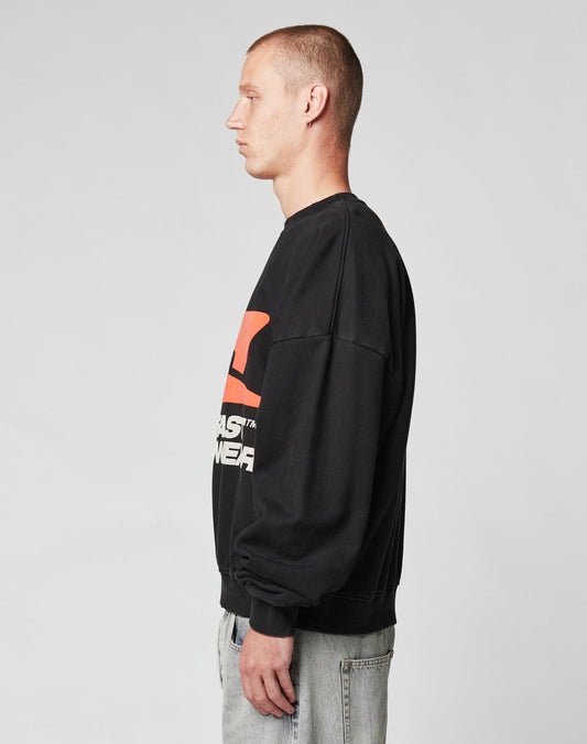 A person with short hair stands in profile against a plain gray background, wearing the LFDY Hardwear Sweater by LIVE FAST DIE YOUNG—an oversized black cotton sweatshirt with a large red pufferprint and white text—paired with light blue jeans.