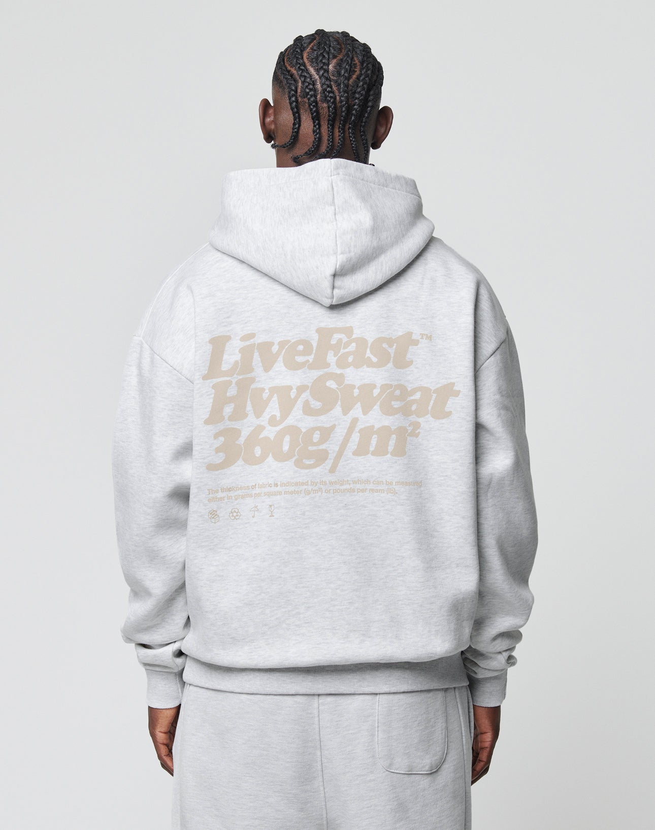 A person with braided hair, seen from behind, wears an oversize light gray Basic 360 Hooded sweatshirt and sweatpants by LIVE FAST DIE YOUNG, featuring large beige Live Fast™ text printed on the back.