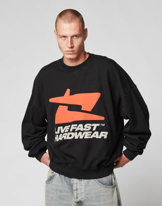 A person with short hair wears the black LFDY Hardwear Sweater by LIVE FAST DIE YOUNG, featuring an orange Pufferprint graphic and white LIVE FAST HARDWEAR text, standing with hands in light blue jean pockets against a plain background.