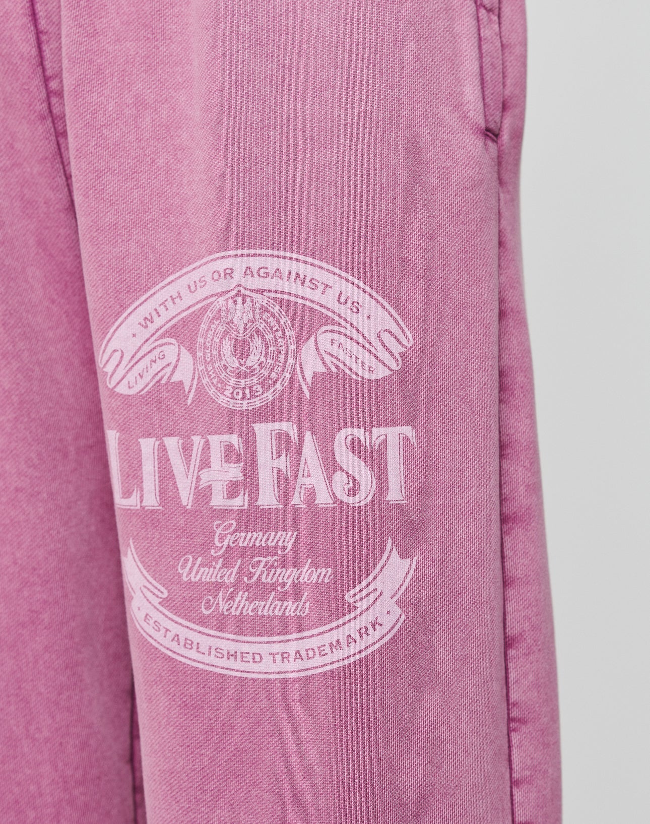 The Loyal Loose Jogger by LIVE FAST DIE YOUNG features pink Baumwolle fabric with white “LIVE FAST” text, banners, a circular emblem, and country names. Oversize fit and vintage wash deliver a bold look.