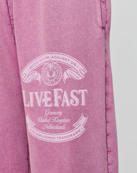 The Loyal Loose Jogger by LIVE FAST DIE YOUNG features pink Baumwolle fabric with white “LIVE FAST” text, banners, a circular emblem, and country names. Oversize fit and vintage wash deliver a bold look.