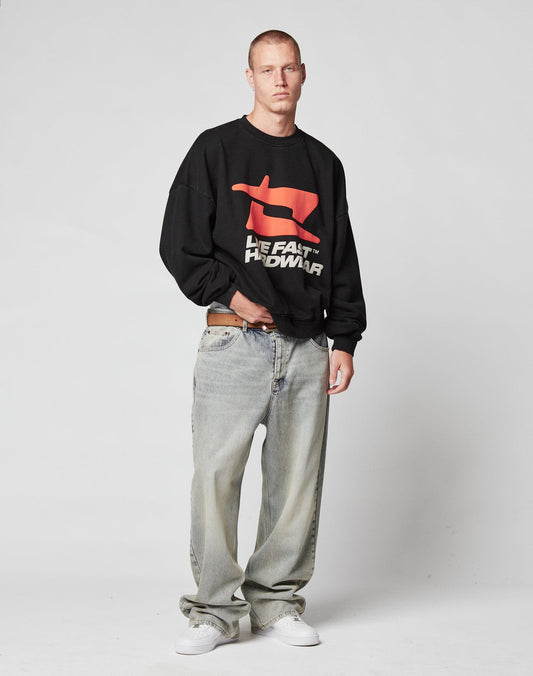 A man with short hair and a neutral expression wears the black LFDY Hardwear Sweater by LIVE FAST DIE YOUNG in Oversize Fit with red and white Pufferprint text, light-washed baggy jeans, and white sneakers. The sweatshirt is made from Baumwolle.