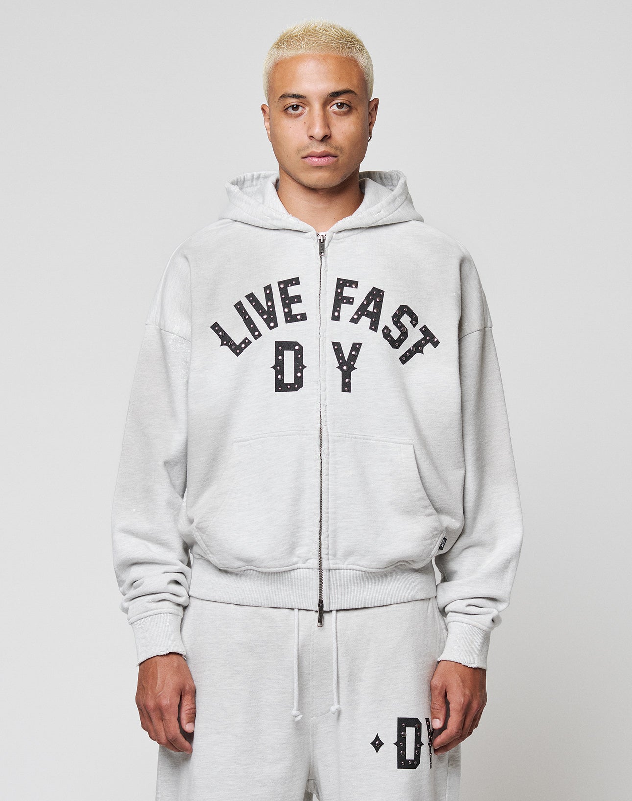 A person with short bleached blonde hair wears the LIVE FAST DIE YOUNG Live Fast Rivet Hooded Zip in light gray, styled oversized with matching sweatpants. Bold black LIVE FAST D*Y text and subtle distressing make it stand out against a white background. | model