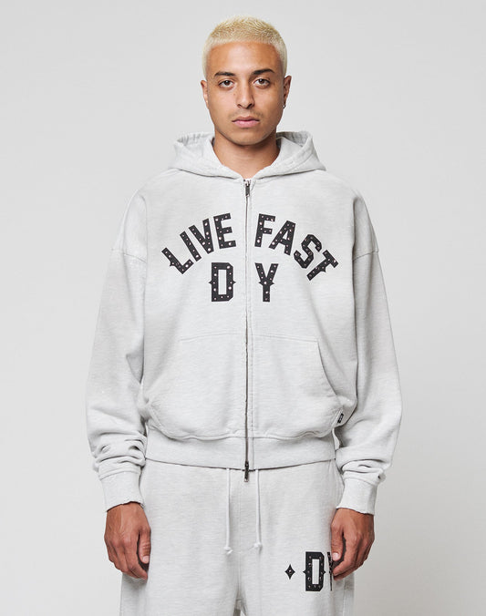 A person with short bleached blonde hair wears the LIVE FAST DIE YOUNG Live Fast Rivet Hooded Zip in light gray, styled oversized with matching sweatpants. Bold black LIVE FAST D*Y text and subtle distressing make it stand out against a white background. | model