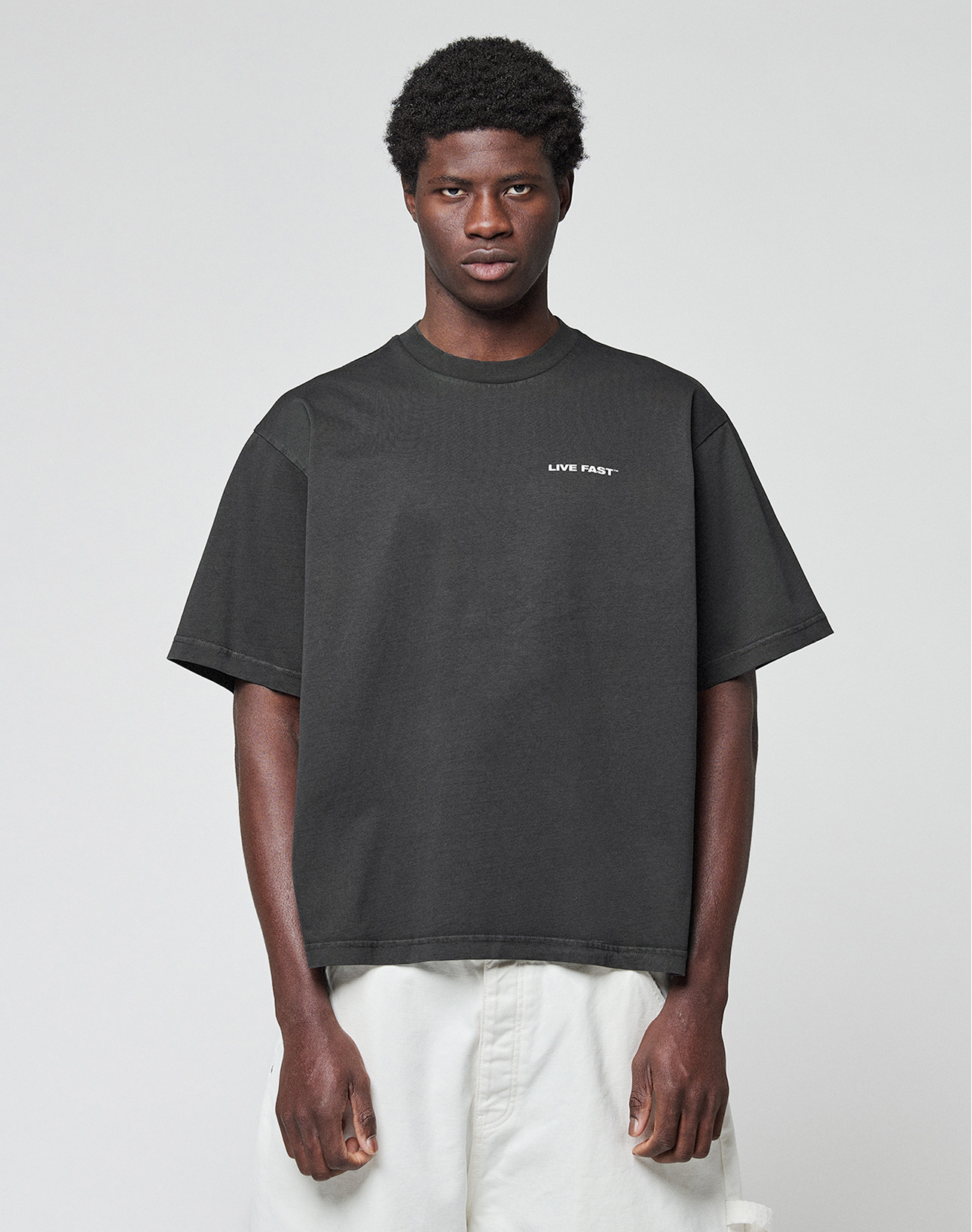 A man wears the LFDY Grand Theft Tee by LIVE FAST DIE YOUNG—an oversized black 100% Baumwolle shirt with "LIVE FAST" on the left chest—paired with white pants, standing against a light gray background and facing the camera.