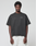 A man wears the LFDY Grand Theft Tee by LIVE FAST DIE YOUNG—an oversized black 100% Baumwolle shirt with "LIVE FAST" on the left chest—paired with white pants, standing against a light gray background and facing the camera.