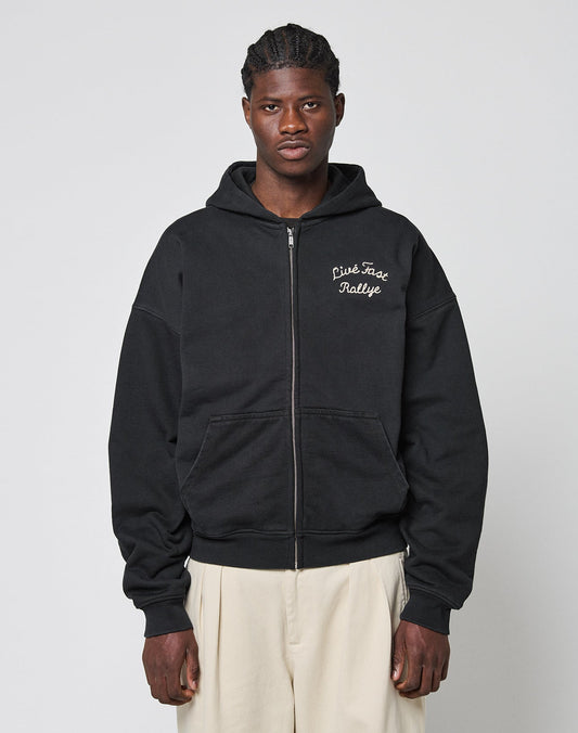 A person wears the LIVE FAST DIE YOUNG LFDY LÉquipe Hooded Zip, an oversized black zip-up hoodie with white cursive chest text, paired with light wide-leg pants, standing against a plain light gray background.
