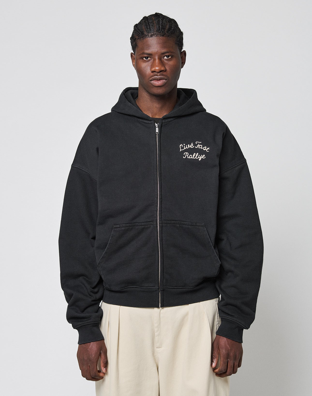A person wears the LIVE FAST DIE YOUNG LFDY LÉquipe Hooded Zip, an oversized black zip-up hoodie with white cursive chest text, paired with light wide-leg pants, standing against a plain light gray background.