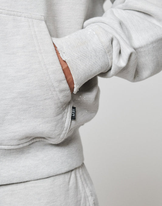 A person wears the LIVE FAST DIE YOUNG Live Fast Rivet Hooded Zip in light gray, with their right hand in the front pocket. The image focuses on the midsection, highlighting a small fabric tag on the sweatshirts pocket.