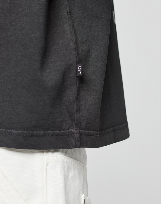 Close-up of the lower side of the LFDY Grand Theft Tee by LIVE FAST DIE YOUNG, featuring a small “FRGMT” fabric tag at the hem, styled with white pants. Focus is on the shirt’s hem and side seam.