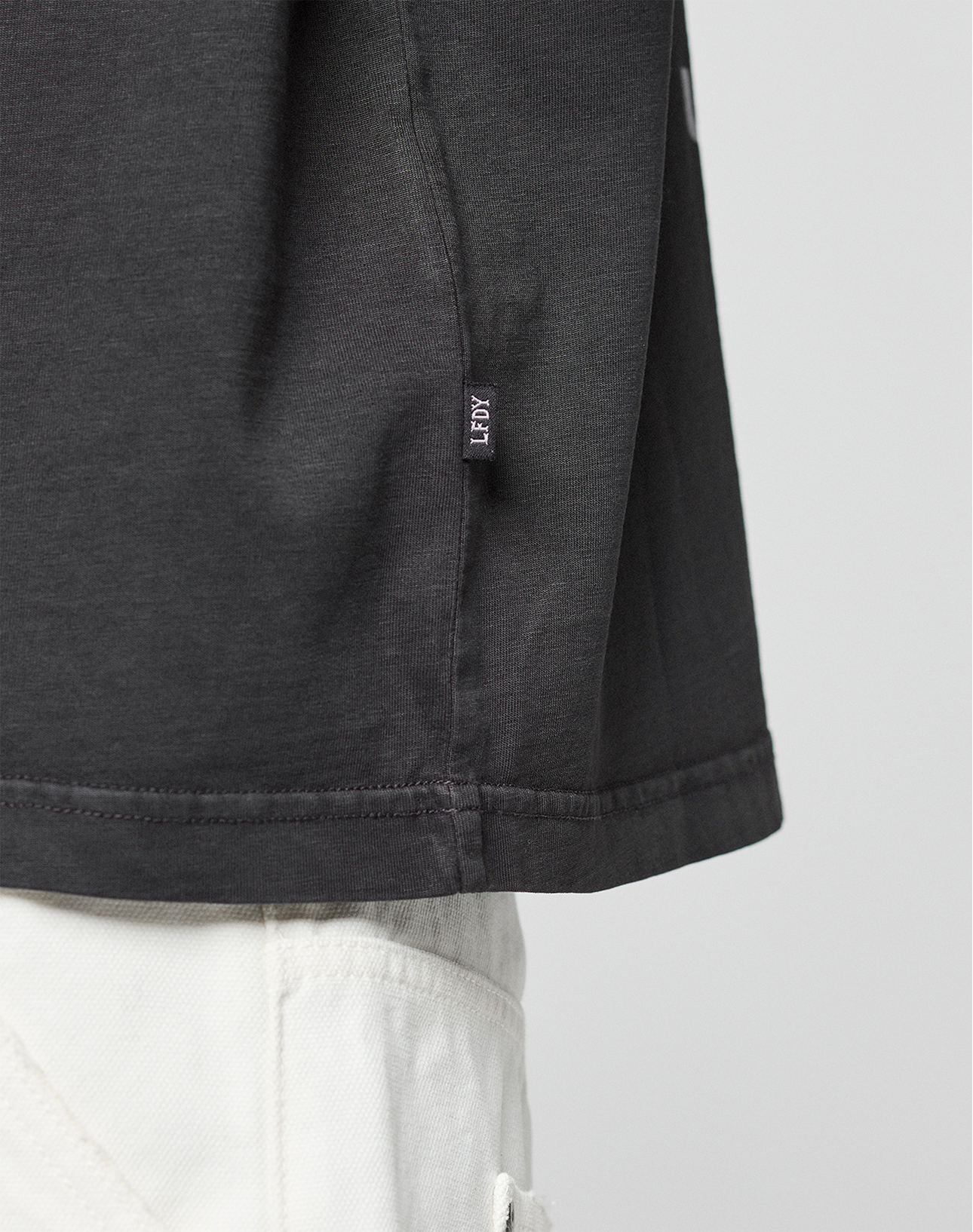 Close-up of the lower side of the LFDY Grand Theft Tee by LIVE FAST DIE YOUNG, featuring a small “FRGMT” fabric tag at the hem, styled with white pants. Focus is on the shirt’s hem and side seam.