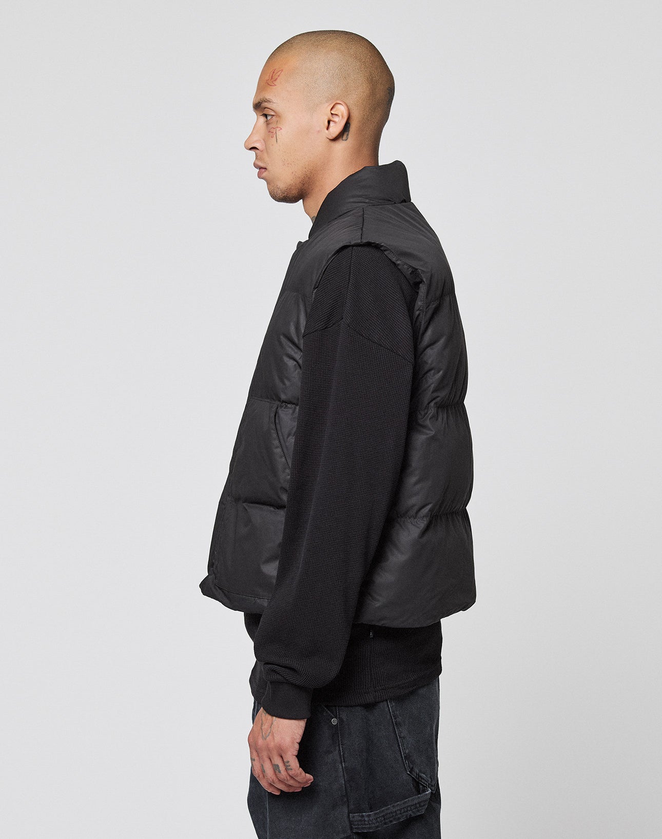 A man with a shaved head stands in profile against a plain background, wearing the LIVE FAST DIE YOUNG Basic Cropped Vest in black over a black long-sleeve shirt and dark jeans.