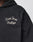 Close-up of the LFDY LÉquipe Hooded Zip by LIVE FAST DIE YOUNG—an oversized black hoodie with Live Fast Rallye embroidered in white chenille on the chest. Part of the persons neck and chin are visible; their face is largely out of frame.