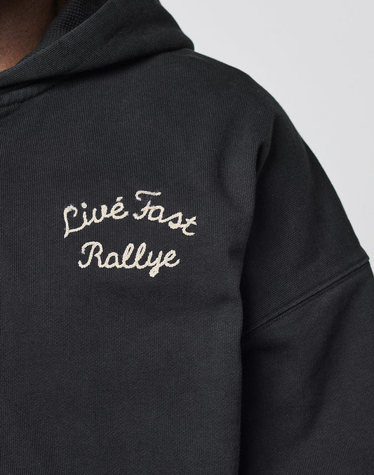 Close-up of the LFDY LÉquipe Hooded Zip by LIVE FAST DIE YOUNG—an oversized black hoodie with Live Fast Rallye embroidered in white chenille on the chest. Part of the persons neck and chin are visible; their face is largely out of frame.