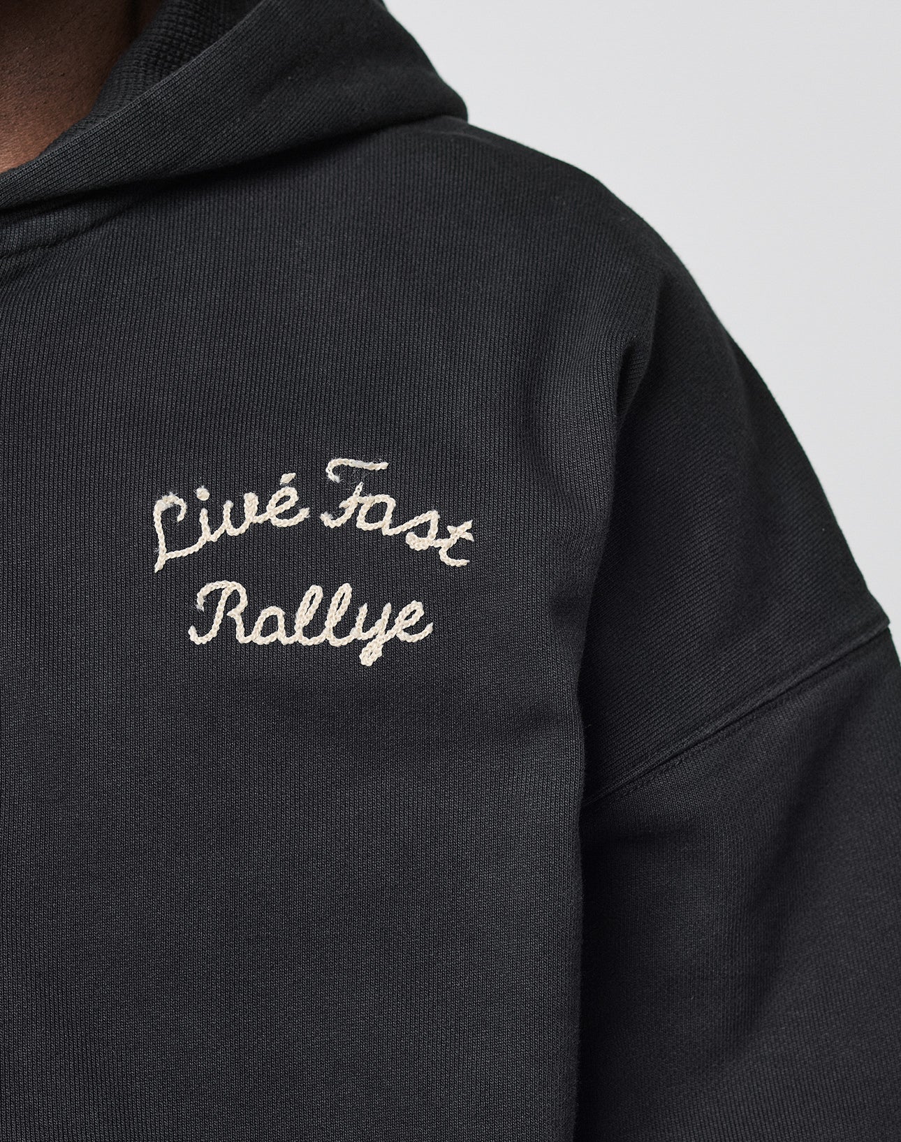 Close-up of the LFDY LÉquipe Hooded Zip by LIVE FAST DIE YOUNG—an oversized black hoodie with Live Fast Rallye embroidered in white chenille on the chest. Part of the persons neck and chin are visible; their face is largely out of frame.