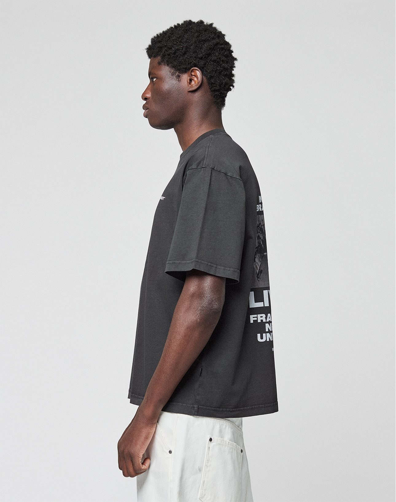 A man with short curly hair stands in profile, wearing the LIVE FAST DIE YOUNG LFDY Grand Theft Tee featuring front and back prints and light-colored shorts against a plain white background.