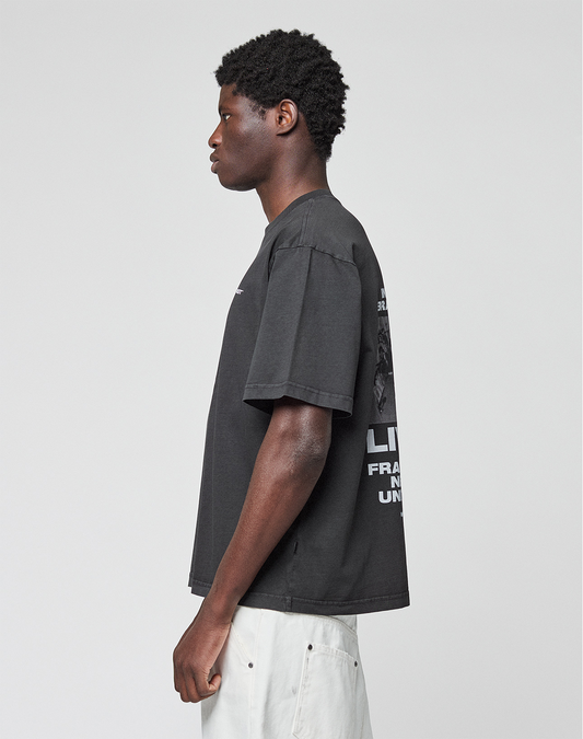 A man with short curly hair stands in profile, wearing the LIVE FAST DIE YOUNG LFDY Grand Theft Tee featuring front and back prints and light-colored shorts against a plain white background.