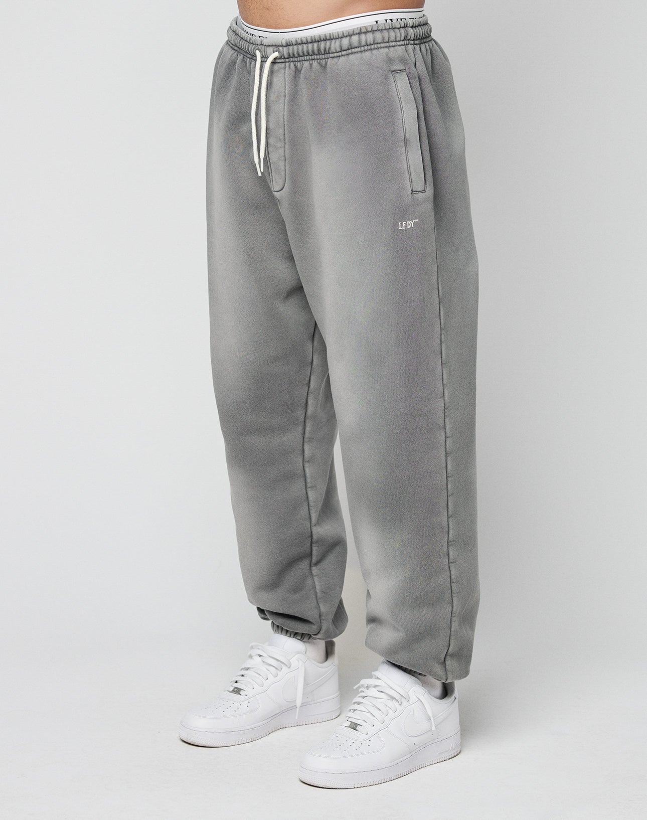 A person models the LIVE FAST DIE YOUNG Basic Loose Jogger—gray, loose-fitting with a faded look, drawstring waist, and side pockets—paired with white sneakers, standing against a plain light background. | model