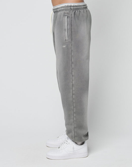 A person stands sideways against a plain light background, wearing LIVE FAST DIE YOUNG’s Basic Loose Jogger in gray with an elastic waistband, white drawstring, subtle logo print, and paired with white sneakers.