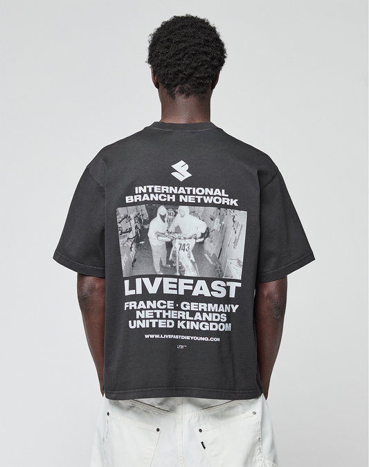 A person with short curly hair, seen from behind, wears the LIVE FAST DIE YOUNG LFDY Grand Theft Tee—an oversize black 100% cotton shirt with front and back prints: “INTERNATIONAL BRANCH NETWORK,” “LIVEFAST,” and various countries. | model