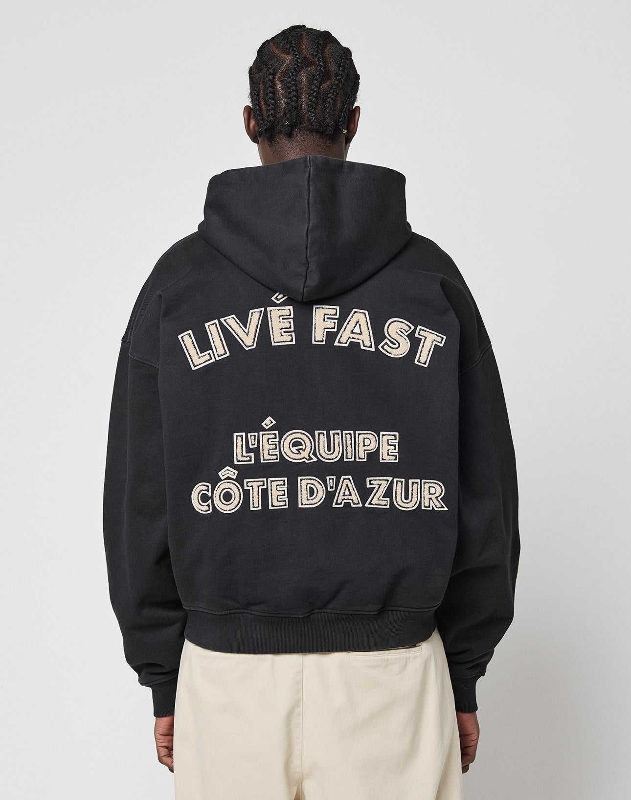 A person with braided hair, seen from behind, wears a black LIVE FAST DIE YOUNG LFDY LÉquipe Hooded Zip with “LIVE FAST L’ÉQUIPE CÔTE D’AZUR” in bold white letters on the back and beige pants. | model