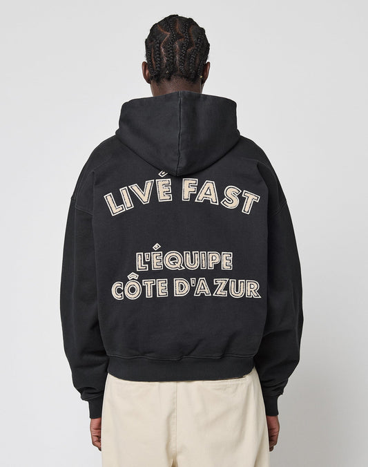 A person with braided hair, seen from behind, wears a black LIVE FAST DIE YOUNG LFDY LÉquipe Hooded Zip with “LIVE FAST L’ÉQUIPE CÔTE D’AZUR” in bold white letters on the back and beige pants. | model