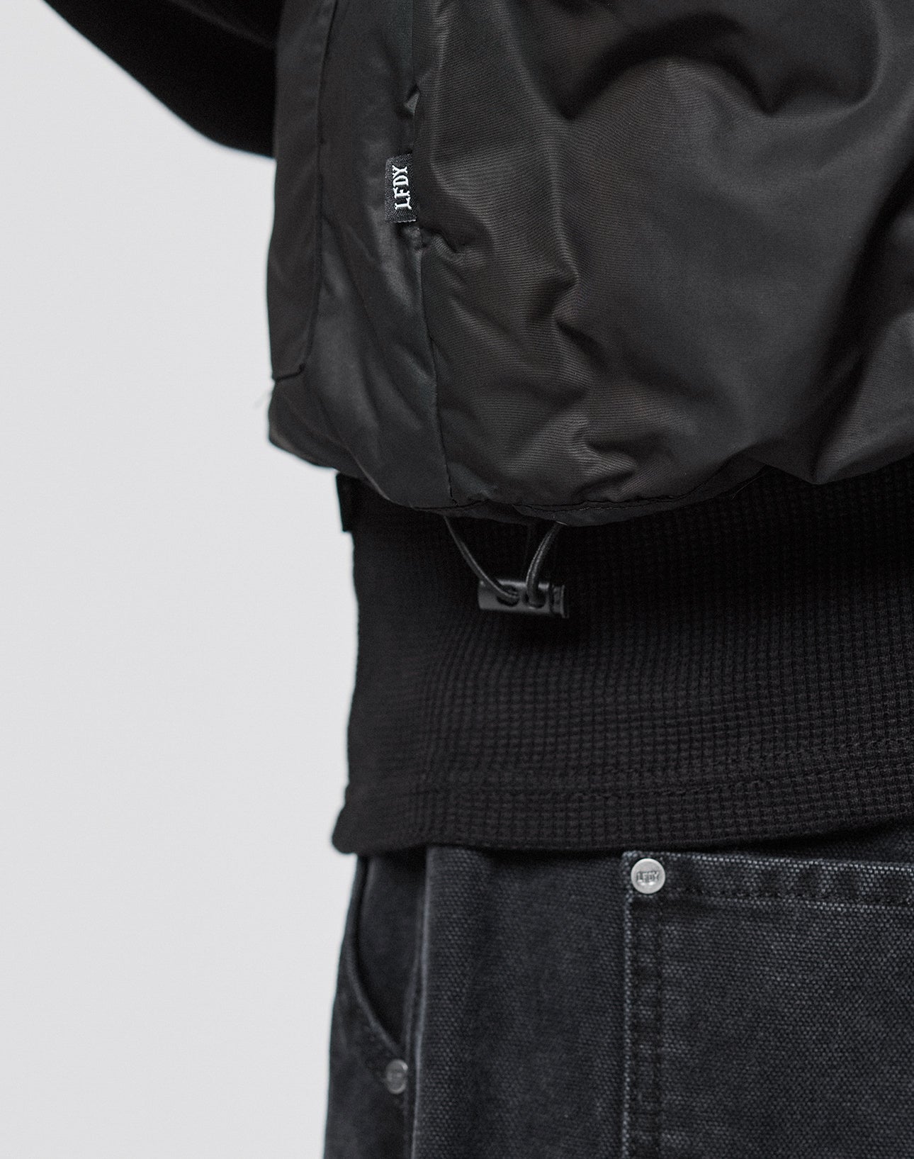 A close-up of a person wearing the LIVE FAST DIE YOUNG Basic Cropped Vest with große Taschen, paired with a textured black sweater and black denim pants featuring silver buttons, focusing on the lower jacket and waist area.