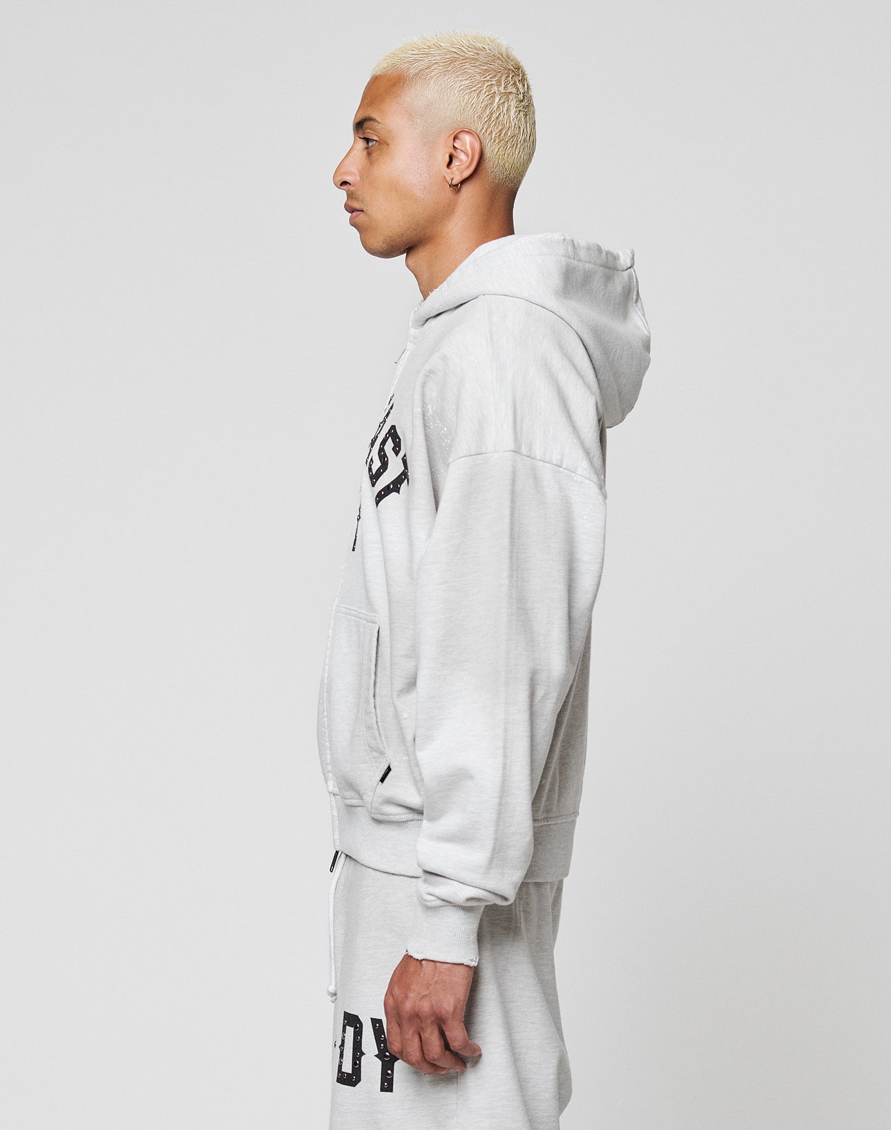 A person with short, bleached blond hair stands in profile, wearing the LIVE FAST DIE YOUNG Live Fast Rivet Hooded Zip sweatshirt in light gray with black lettering and handgefertigter Sprayeffekt, against a plain white background.