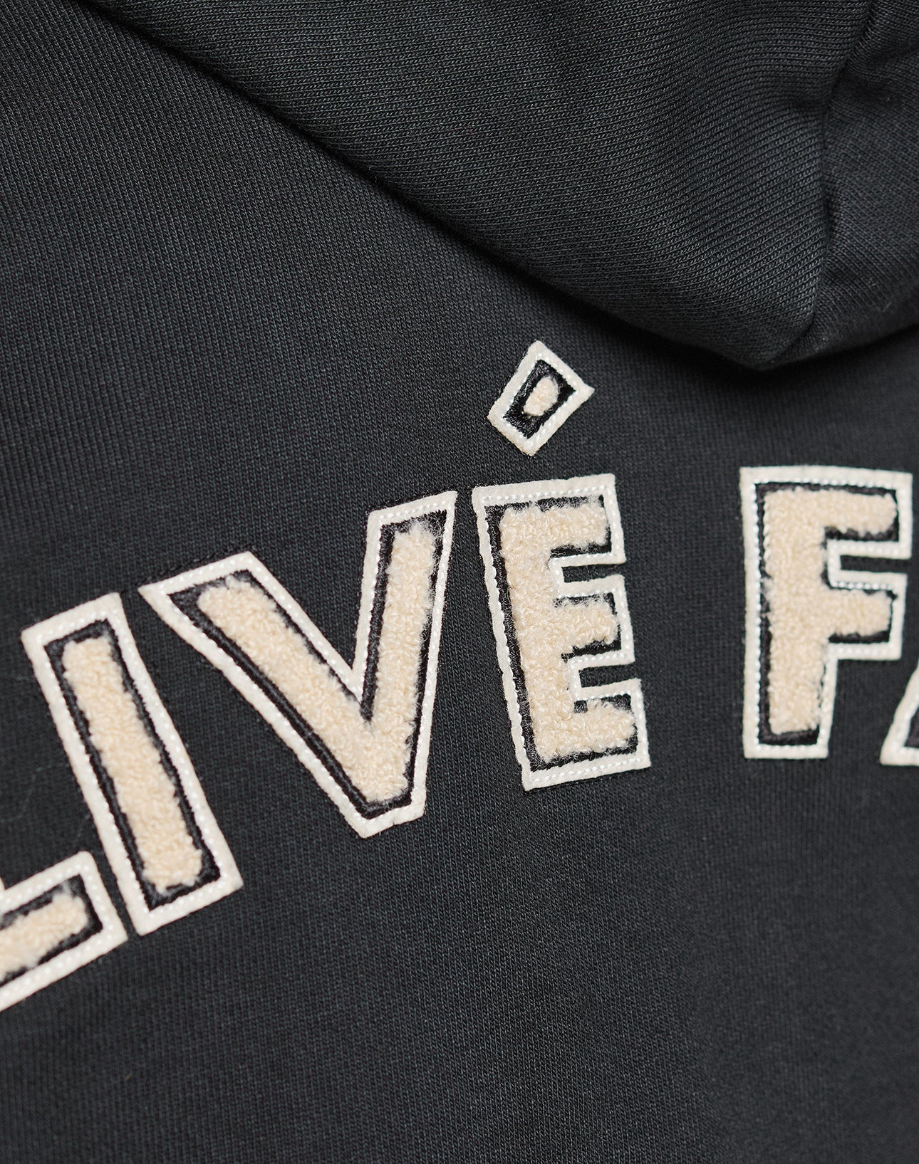 Close-up of the black LFDY LÉquipe Hooded Zip by LIVE FAST DIE YOUNG, showing raised stitched white letters and bold chenille stick-outlined embroidery for standout texture.