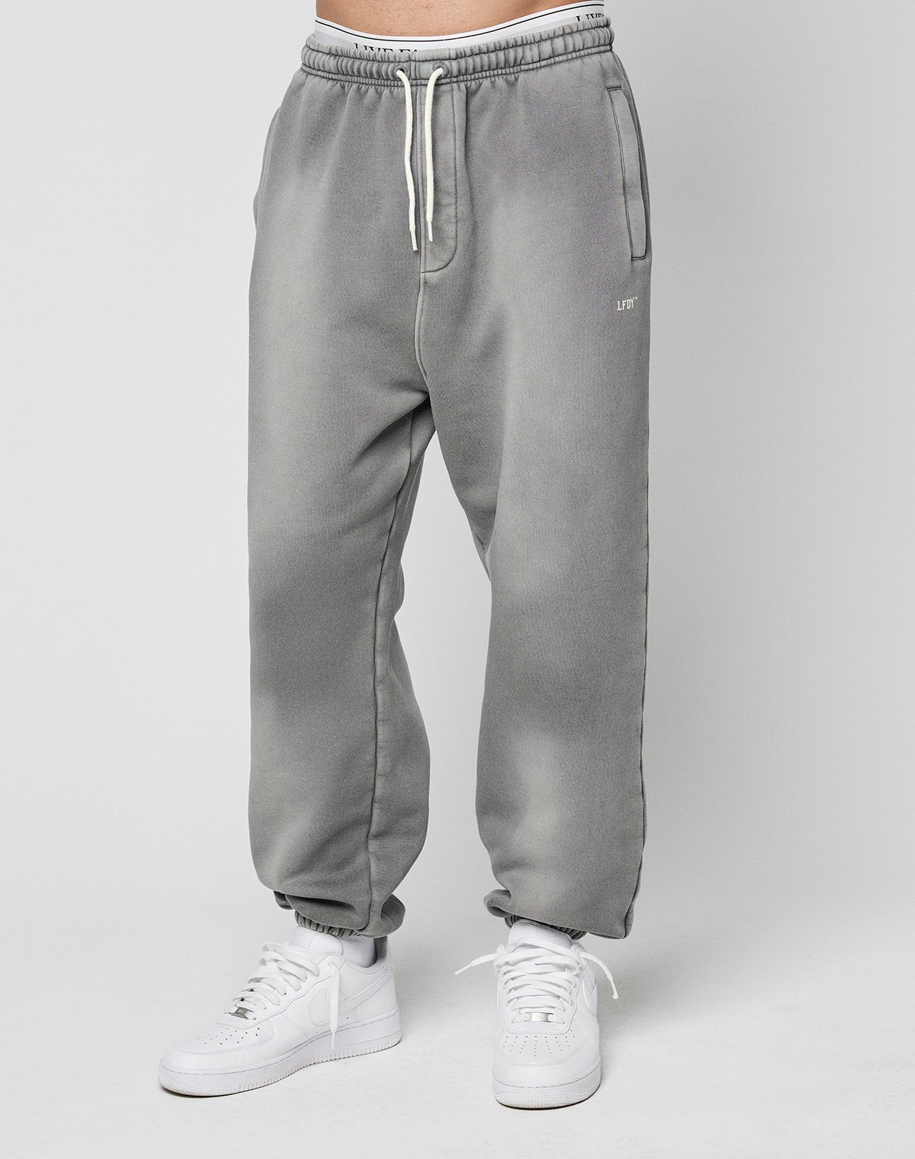 A person models the LIVE FAST DIE YOUNG Basic Loose Jogger—light gray, loose-fitting sweatpants with a logo print and white drawstrings—paired with white sneakers against a plain light gray background.